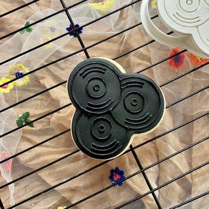 May include: A black cookie shaped like three vinyl records, with concentric circles, sits on a cooling rack. A white cookie cutter in the shape of the cookie is in the upper right corner. The rack is over a beige surface with colorful floral embroidery.