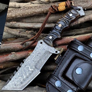 May include: A Damascus steel knife with a dark, textured handle and a leather lanyard. The blade has a unique pattern and a serrated edge. A black leather sheath with silver-colored rivets is also visible.