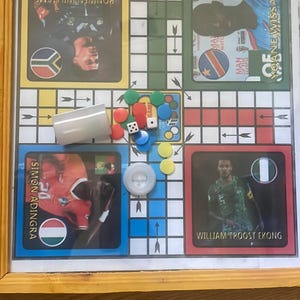 Pu&ograve; includere: Un gioco da tavolo Ludo incorniciato con carte giocatore con calciatori. Include dadi, pedine e un bicchiere per i dadi. Il tabellone &egrave; su una superficie di legno.