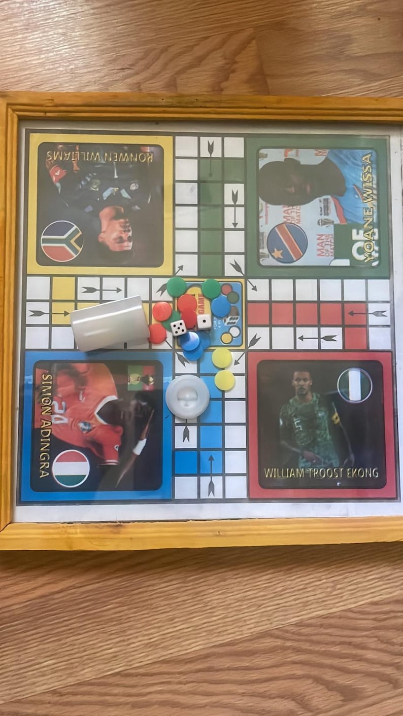 Pu&ograve; includere: Un gioco da tavolo Ludo incorniciato con carte giocatore con calciatori. Include dadi, pedine e un bicchiere per i dadi. Il tabellone &egrave; su una superficie di legno.