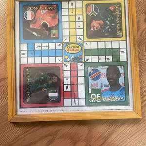 Pu&ograve; includere: Un gioco da tavolo Ludo incorniciato con una cornice di legno gialla. Il tabellone presenta ritratti di giocatori e bandiere nazionali. Il centro del tabellone riporta il testo "Original Ludo Game".