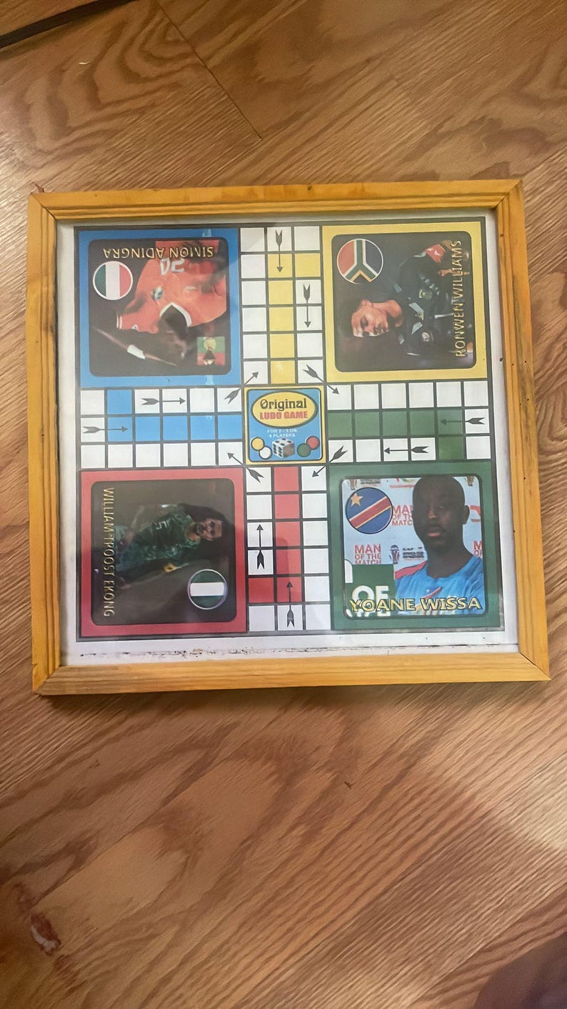 Pu&ograve; includere: Un gioco da tavolo Ludo incorniciato con una cornice di legno gialla. Il tabellone presenta ritratti di giocatori e bandiere nazionali. Il centro del tabellone riporta il testo "Original Ludo Game".