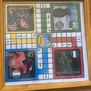 Pu&ograve; includere: Gioco del Ludo incorniciato con ritratti di giocatori e bandiere nazionali. Il tabellone ha un bordo giallo e include il testo "Original Ludo Game". I giocatori sono Simon Adingra, Ronwen Williams, Yoane Wissa e William Troost-Ekong.