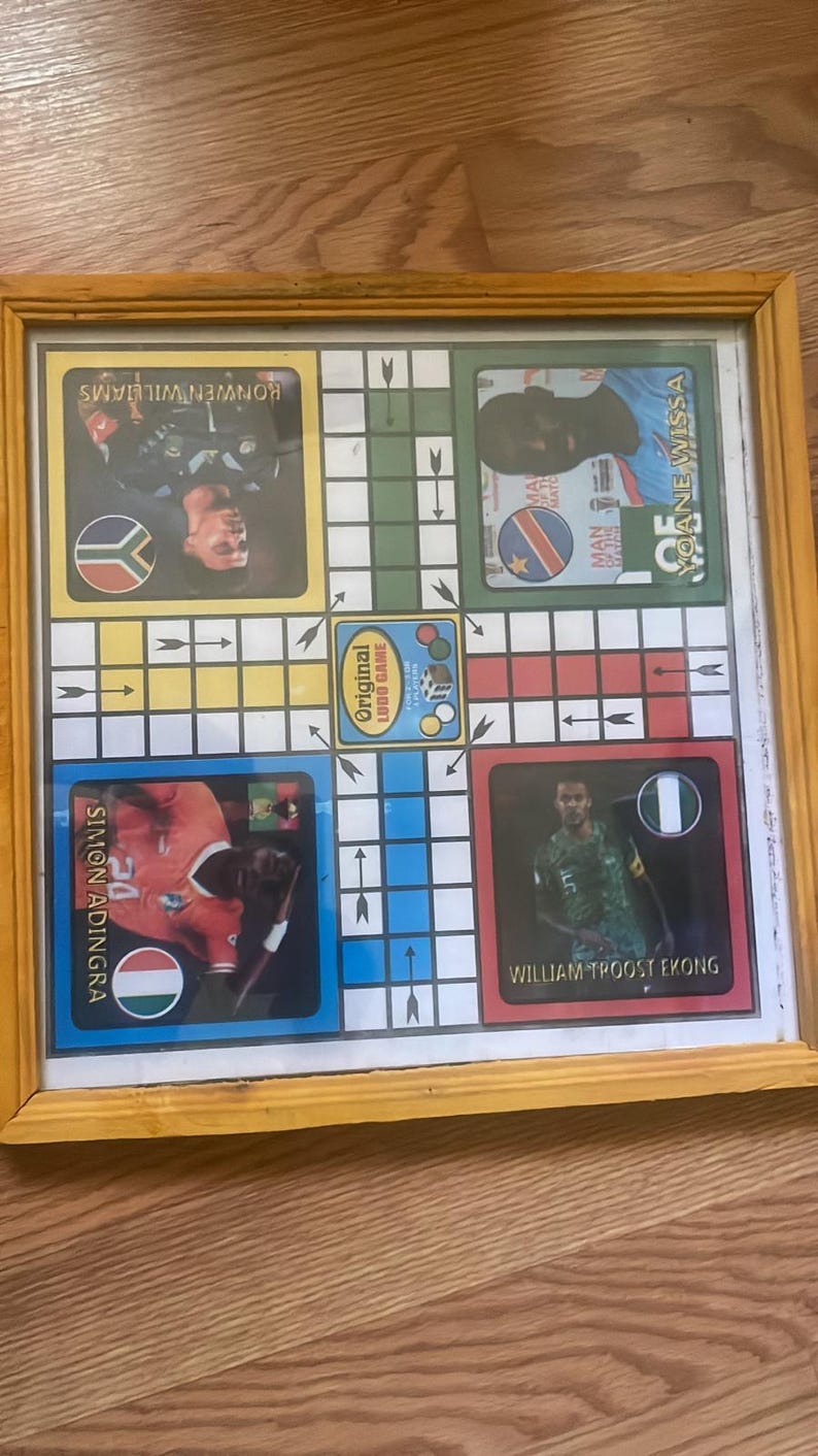 Pu&ograve; includere: Gioco del Ludo incorniciato con ritratti di giocatori e bandiere nazionali. Il tabellone ha un bordo giallo e include il testo "Original Ludo Game". I giocatori sono Simon Adingra, Ronwen Williams, Yoane Wissa e William Troost-Ekong.