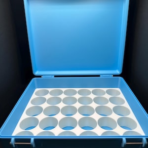 May include: Open, light blue plastic carrying case with a white interior insert featuring multiple circular compartments. The case has a hinged lid and a latch closure. The interior insert is designed to hold small items.