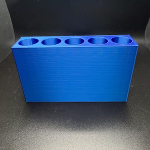 May include: A bright blue, rectangular organizer with five circular openings. The organizer is made of a solid material and has a textured surface. The item is designed to hold small cylindrical objects.