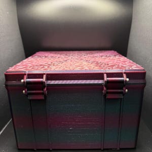 May include: A rectangular storage box with a geometric patterned lid. The box is primarily dark with a gradient of red and green hues. The lid is a lighter shade of red with a geometric pattern. The box has two metal hinges and a latch.