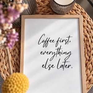 May include: A rectangular wooden frame containing a white print with the black text Coffee first, everything else later. The frame is positioned on a woven circular mat, accompanied by a glass candle holder and dried purple flowers.