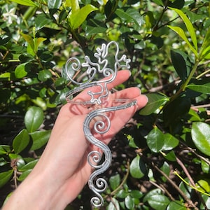 May include: A silver wire arm cuff with a lizard design. The cuff has a decorative, swirling pattern and a lizard-shaped accent. The jewellery is displayed against a backdrop of green foliage.