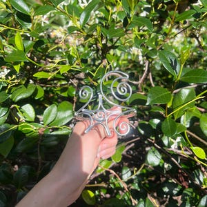 May include: A silver wire bracelet featuring a starburst design at its centre, with spiral embellishments. The bracelet is displayed against a background of green leaves and small white flowers.