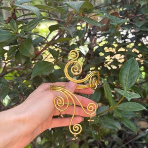May include: Gold-coloured arm cuff with a swirling design. The cuff features multiple spiral accents and small star-shaped charms. The jewellery is held against a backdrop of green foliage.