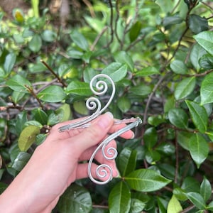 May include: Two silver-colored hair accessories with a swirling design are held in front of a green leafy background. The accessories have a delicate, wire-like appearance with spiral embellishments.