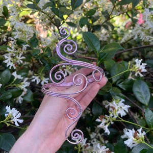 Könnte beinhalten: Ein zartes, lavendelfarbenes Armband mit einem wirbelnden Drahtdesign. Das Armband hat mehrere Spiralakzente und ist vor einem Hintergrund aus grünen Blättern und weißen Blüten angeordnet.