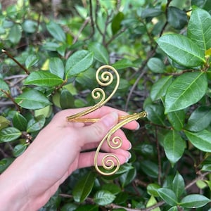 May include: A gold arm cuff with a decorative swirl design. The cuff is held against a backdrop of green foliage. The cuff features two spiral designs and is made of thin, curved metal.