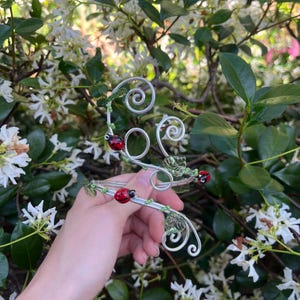 Può includere: Un bracciale in filo metallico color argento con tre piccoli ciondoli a forma di coccinella rossa e nera, arricchito da foglie in vetro verde traslucido e delicati dettagli in perline verdi. Il filo forma diverse spirali circolari.