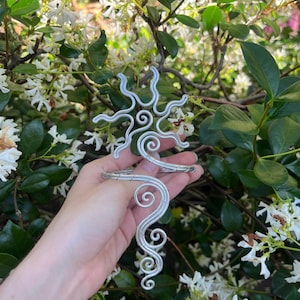 Könnte beinhalten: Ein Oberarmreif aus silberfarbenem Draht mit einem sonnenförmigen Design an der Oberseite, das strahlende, wellenförmige Strahlen und ein nach unten verlaufendes Spiralmuster aufweist. Der Draht ist zu komplexen, symmetrischen Wirbeln geformt.