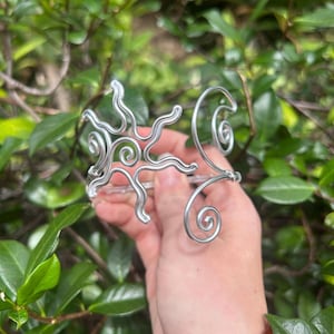 Könnte beinhalten: Ein silberfarbenes Draht-Armreif-Armband mit einem Sonnenstrahlen-Design. Das Armband hat wirbelnde, spiralförmige und wellenförmige Drahtelemente. Der Schmuck wird vor einem Hintergrund aus grünem Laub gehalten.