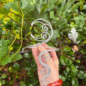 Pode incluir: Uma pulseira de braço e um adorno de cabeça de arame prateado com um design de lua crescente e detalhes em espiral. A pulseira é usada com uma pulseira vermelho-alaranjada. O fundo apresenta folhagem verde.