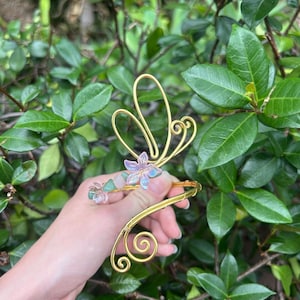 Könnte beinhalten: Ein goldfarbenes Draht-Manschettenarmband mit Schmetterlingsdesign. Das Armband hat einen floralen Akzent mit lila und klaren Kristallen. Das Armband wird vor einem Hintergrund aus grünen Blättern gehalten.