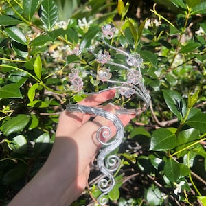 May include: A silver-coloured, handcrafted headpiece with a floral design. The headpiece features delicate, wire-wrapped branches adorned with small, pink and clear flower accents. The design includes a curved, swirling base. The background is lush green foliage.