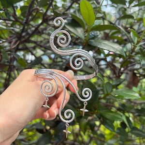 May include: Silver arm cuff with a decorative, swirling wire design. The cuff features multiple spiral accents and small cross-shaped charms hanging from the spirals. The jewelry is displayed against a backdrop of green foliage.