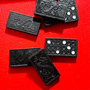 Puede incluir: Domin&oacute;s negros con puntos blancos y relieves de dragones. Los domin&oacute;s est&aacute;n dispersos sobre una superficie roja. Algunos domin&oacute;s tienen puntos blancos, otros dise&ntilde;os detallados de dragones.