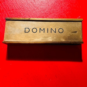 Puede incluir: Una caja de domin&oacute; de madera con la palabra "DOMINO" impresa en negro. La caja es rectangular y tiene un acabado marr&oacute;n claro. El fondo es rojo liso.