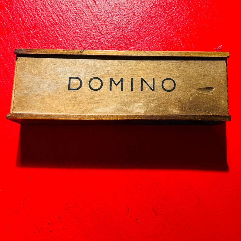 Puede incluir: Una caja de domin&oacute; de madera con la palabra "DOMINO" impresa en negro. La caja es rectangular y tiene un acabado marr&oacute;n claro. El fondo es rojo liso.