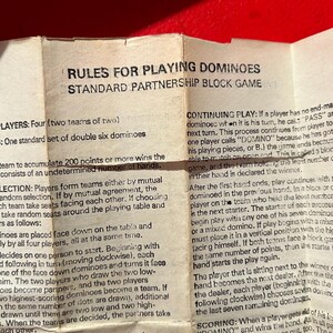 Puede incluir: Primer plano de un papel doblado con reglas para jugar al domin&oacute; en asociaci&oacute;n. El texto negro est&aacute; sobre un fondo beige. El papel est&aacute; sobre una superficie roja. Instrucciones de juego y puntuaci&oacute;n.