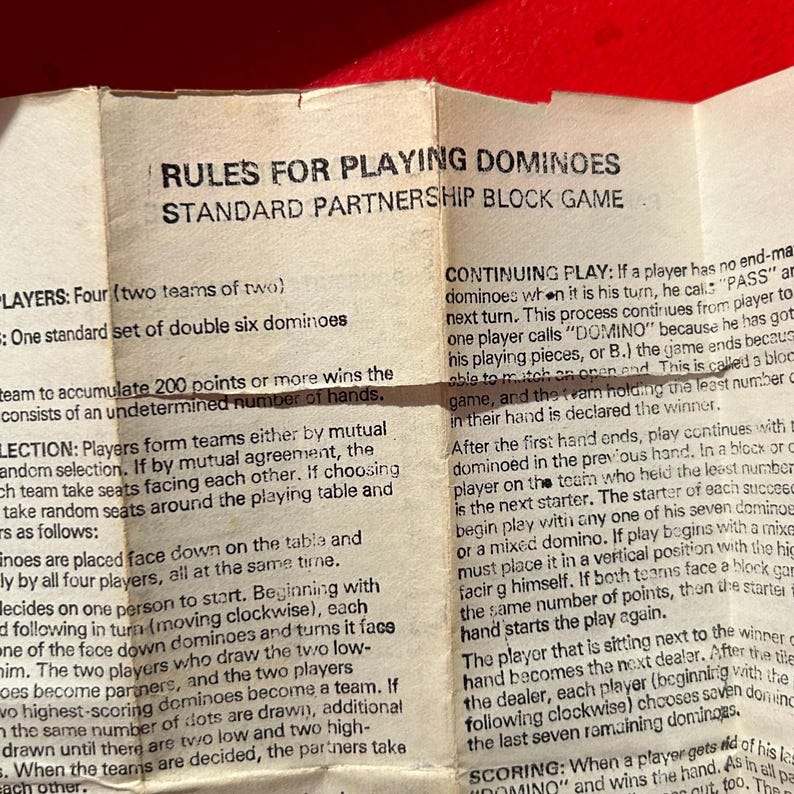 Puede incluir: Primer plano de un papel doblado con reglas para jugar al domin&oacute; en asociaci&oacute;n. El texto negro est&aacute; sobre un fondo beige. El papel est&aacute; sobre una superficie roja. Instrucciones de juego y puntuaci&oacute;n.