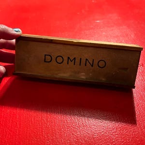 Puede incluir: Una caja de domin&oacute; de madera con la palabra "DOMINO" impresa en negro. La caja es rectangular y tiene un aspecto vintage desgastado. El fondo es de color rojo liso.
