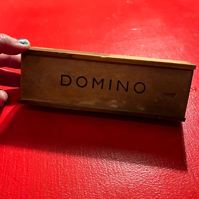 Puede incluir: Una caja de domin&oacute; de madera con la palabra "DOMINO" impresa en negro. La caja es rectangular y tiene un aspecto vintage desgastado. El fondo es de color rojo liso.