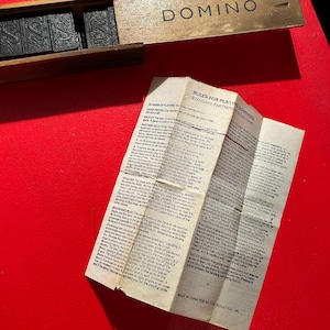 Puede incluir: Un juego de domin&oacute; en una caja de madera con la palabra "DOMINO" impresa en la tapa. El juego incluye fichas de domin&oacute; negras con marcas blancas. Tambi&eacute;n hay un papel doblado con las reglas del juego.