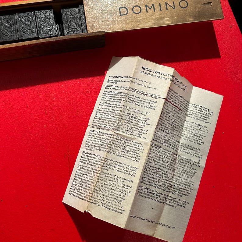 Puede incluir: Un juego de domin&oacute; en una caja de madera con la palabra "DOMINO" impresa en la tapa. El juego incluye fichas de domin&oacute; negras con marcas blancas. Tambi&eacute;n hay un papel doblado con las reglas del juego.