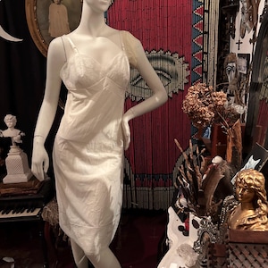 May include: A white slip dress with thin straps and a lace trim. The dress is on a mannequin in a room with a variety of decorative items.