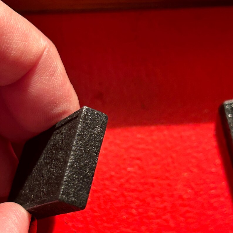 Puede incluir: Primer plano de una pieza de juego rectangular negra con una superficie texturizada. La pieza est&aacute; sostenida por una mano sobre un fondo rojo. La pieza del juego parece ser parte de un juego de mesa.