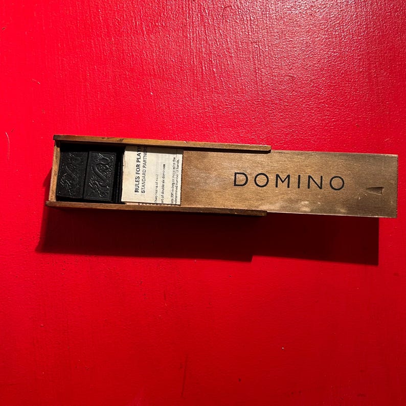Puede incluir: Un juego de domin&oacute; de madera con fichas negras dentro de una caja de madera. La caja est&aacute; abierta, revelando las fichas de domin&oacute; y un papel impreso con reglas. La palabra "DOMINO" est&aacute; impresa en la caja. El fondo es rojo liso.