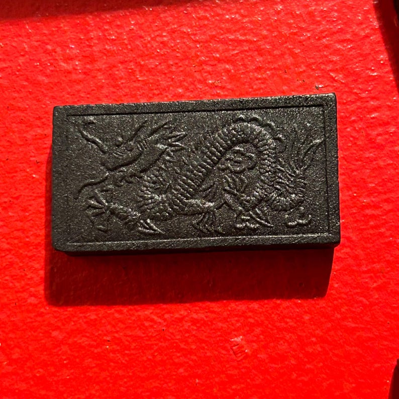 Puede incluir: Una baldosa decorativa rectangular negra con un relieve detallado de un drag&oacute;n. El drag&oacute;n est&aacute; representado en un estilo asi&aacute;tico tradicional, con escamas intrincadas y un cuerpo fluido. La baldosa est&aacute; sobre un fondo rojo.
