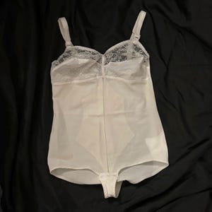 May include: White lace-trimmed bodysuit with a smooth, silky texture. The bodysuit has a zipper closure in the front and a hook and eye closure at the bottom.