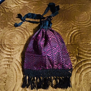 May include: A drawstring bag with a patterned design in shades of purple and black. The bag has a black fringe along the bottom and black ribbons for closure. The bag is sitting on a brown, quilted surface.