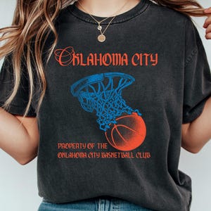 Op de afbeelding: Donkergrijze t-shirt met een basketbalafbeelding. Het ontwerp bevat een basketbal die door een net gaat, met de woorden "Oklahoma City" en "Property of the Oklahoma City Basketball Club" in rood en blauw.