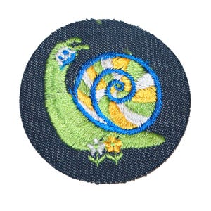 May include: Embroidered patch featuring a whimsical snail design. The snail is green with a spiraling shell in shades of blue, yellow, and white. The patch is circular, set against a dark blue fabric background, and includes small floral accents.