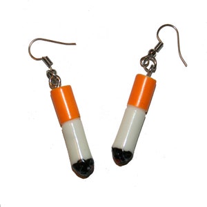 May include: A pair of dangle earrings shaped like cigarettes. The earrings are made of white and orange plastic with a black detail at the end. The earrings have silver hooks.