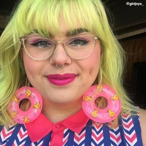 Puede incluir: Una persona con el pelo verde brillante y labios rosas lleva grandes pendientes de aro inflables de color rosa con dise&ntilde;os de patos amarillos. Lleva una camisa con estampado de chevron rosa, blanco y azul.