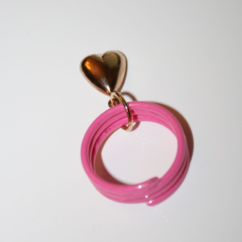 May include: A pink ring with a gold heart charm. The ring is made of multiple layers of pink plastic.