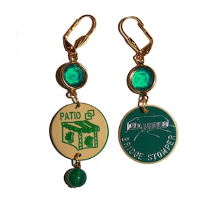 May include: A pair of gold earrings with green gem accents. The earrings feature round, green, glass stones and a charm with a green and beige design. The charm on the left earring says "Patio" and the charm on the right earring says "Bridge Stomper".