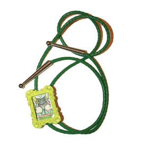 May include: A green bolo tie with a decorative lime green frame pendant. The pendant features a cartoon image and the text "ALOE TURTLES". The tie has silver-colored metal tips and a braided green cord.