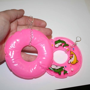 Puede incluir: Un par de pendientes de donut hinchables de color rosa con cadenas plateadas. Un donut tiene un dise&ntilde;o de delf&iacute;n blanco, verde y amarillo con chispas de colores.