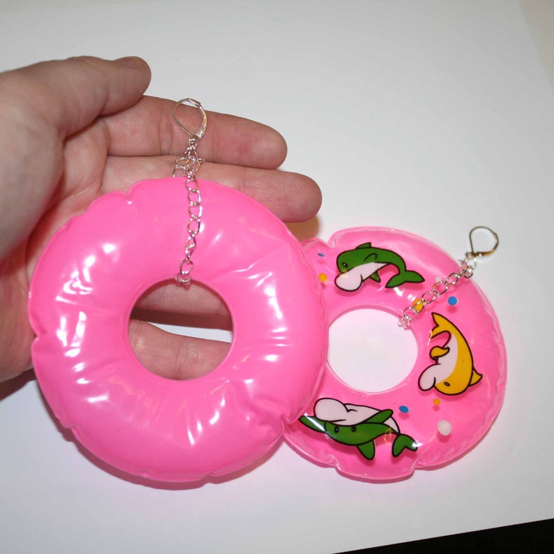 Puede incluir: Un par de pendientes de donut hinchables de color rosa con cadenas plateadas. Un donut tiene un dise&ntilde;o de delf&iacute;n blanco, verde y amarillo con chispas de colores.