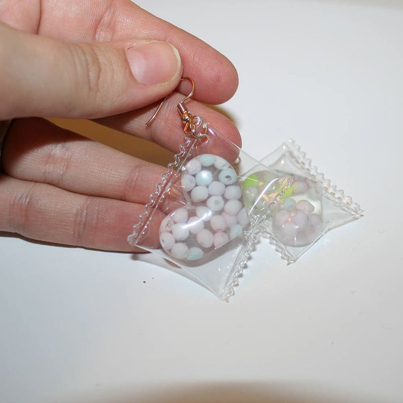 May include: A pair of dangle earrings with a heart-shaped charm filled with pastel-colored beads. The charms are encased in clear plastic that resembles a small candy wrapper.